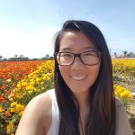 [photo of a girl with long black hair, wearing glasses and white tank top, smiling with orange and yellow flowers behind her and clear blue sky