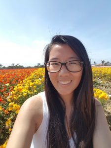 [photo of a girl with long black hair, wearing glasses and white tank top, smiling with orange and yellow flowers behind her and clear blue sky