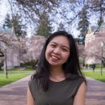 Image description: a photo of Lorie she has short dark brown hair wearing a green dress. Her head is tilted to the right smiling with her hands in front of her. She is standing behind pink cherry blossoms, green grass, and sidewalk bricks in the background.