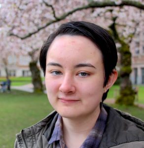 A person is smiling softly towards the camera, with brown jacket and a floral tree in the background.