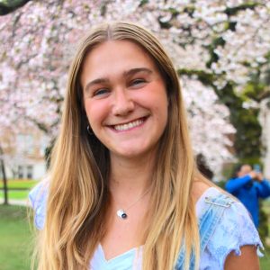 A feminine looking person smiling forwards, wearing white top with blue overalls and floral tree is in the background