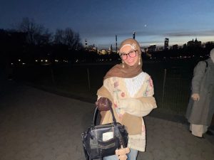  Picture of a smiling Rawan in Central Park. It is dark outside. In the distance there is a dark sky, lit up skyscrapers, and a fenced in grassy area. Rawan is wearing a light brown hijab, black square glasses, and pearl earrings. She has on a yellow, floral knit cardigan layered over a knit white sweater, and blue jeans. She had on brown gloves and carries a black purse in her right hand.